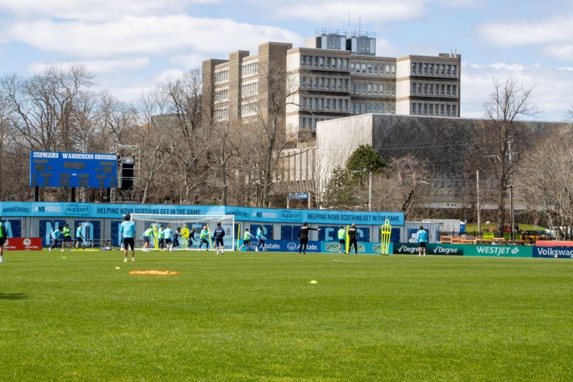 Soccer players practice on a soccer field with a hospital in the background. 