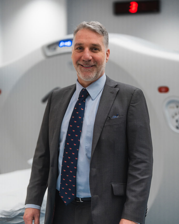 A man in a suit stands in front of a CT scanner in a clinical setting. 