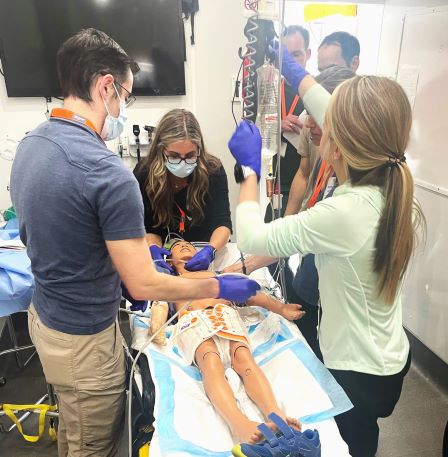 Three people in a healthcare setting are working on a mannequin patient as part of a simulation. The mannequin is laying on a stretcher.