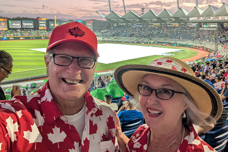 A QEII patient and her partner, dressed in Canadian maple leaf clothing, stand in the crowd overlooking a baseball diamond.