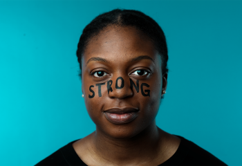 Close up of woman in a black shirt standing in front of a blue background with the words "strong" painted across her face in black text