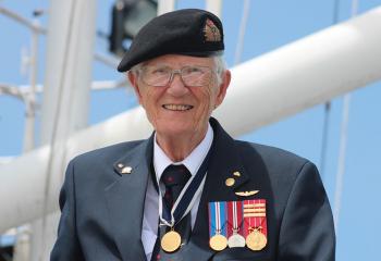 Older woman wearing her  Lieutenant-Commander uniform, stands in front of a white mast belonging to the HMCS Sackville ship