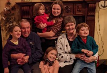 An elderly couple sitting in their living room, surrounded by five of their young grandchildren. Some of the grandchildren are in their laps, all wearing big, bright smiles