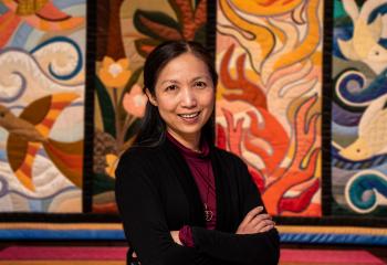 A woman, standing with her arms crossed, smiles at the camera. She stands in front of a colourful tapestry. 