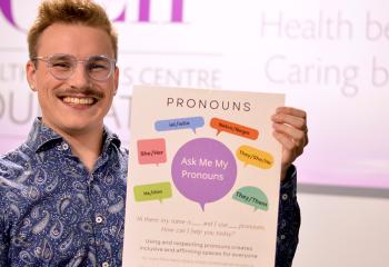 Garry Dart wears a blue patterned shirt and smiles holding up a pronouns poster