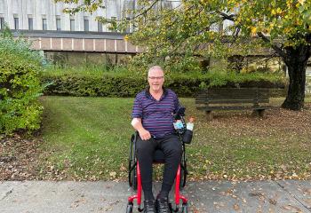 A man sits in a wheelchair in front of a brick building, surrounded by foliage. 