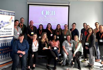 A group of people are posed smiling and facing the camera. A screen is behind them with the QE 2 Foundation logo. 