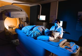 A female patient is laying down, preparing to undergo a medical scan. A female healthcare professional stands close to the patient. 