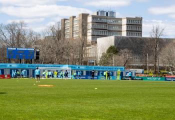 Soccer players practice on a soccer field with a hospital in the background. 