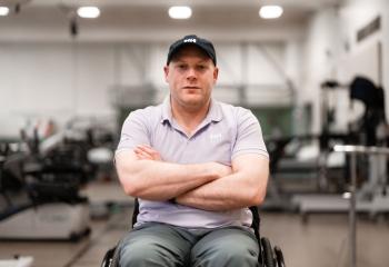 A man sitting in a wheelchair, arms crossed and smiling towards the camera. He is sitting in a gym-type setting. 