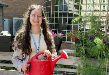 A woman with long brown hair stands smiling outdoors in a garden. In the background is a building with lots of windows. She is surrounded by flowers and plants and holds a watering can.