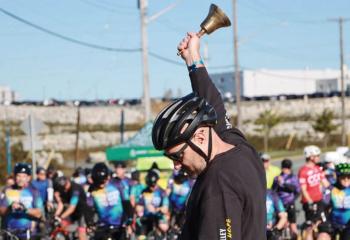A rider wearing a black shirt and helmet rings a bell in front of a large crowd