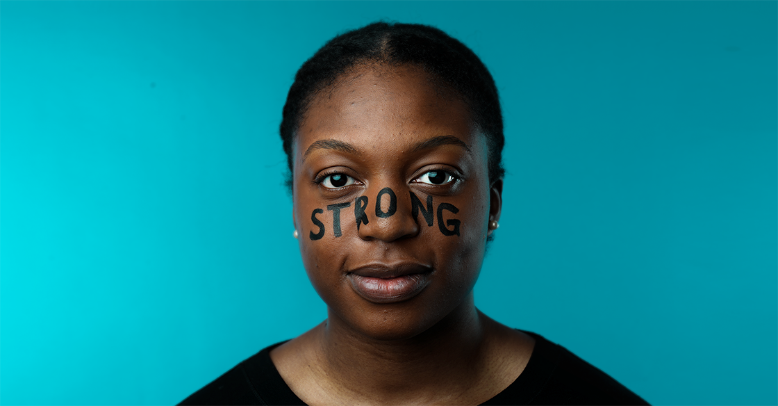 Close up of woman in a black shirt standing in front of a blue background with the words "strong" painted across her face in black text