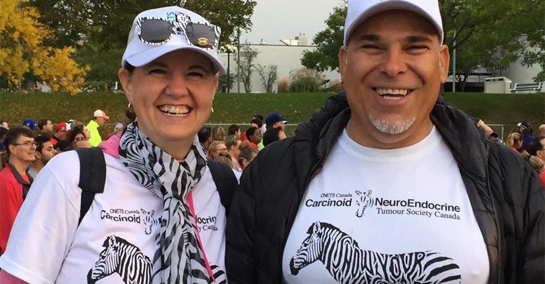 Jackie stands alongside her husband, Joe, at a busy, outdoor event. Both are smiling and wearing white t-shirts with zebra graphics