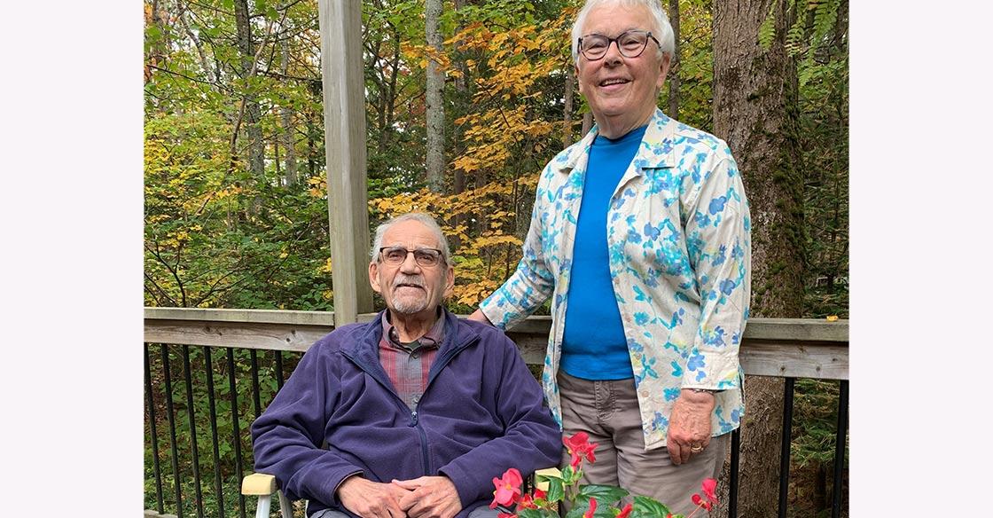 An elderly couple stands on a porch, surrounded by fall colours. The husband, Bob, sits in a chair on the left to his wife, Wendy, who's standing next to him.