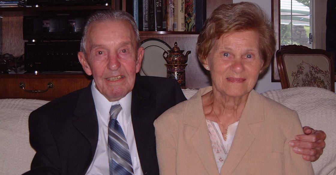 Elderly couple sit together on white sofa, husband's arm is on his wife's shoulder