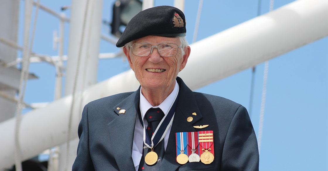 Older woman wearing her Lieutenant-Commander uniform, stands in front of a white mast belonging to the HMCS Sackville ship