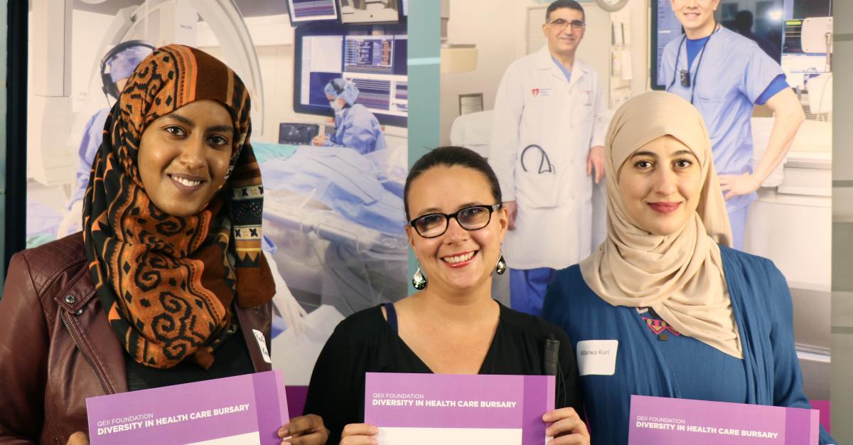 Three women stand against a healthcare backdrop holding QEII Foundation Diversity in Health Care Bursary certificates