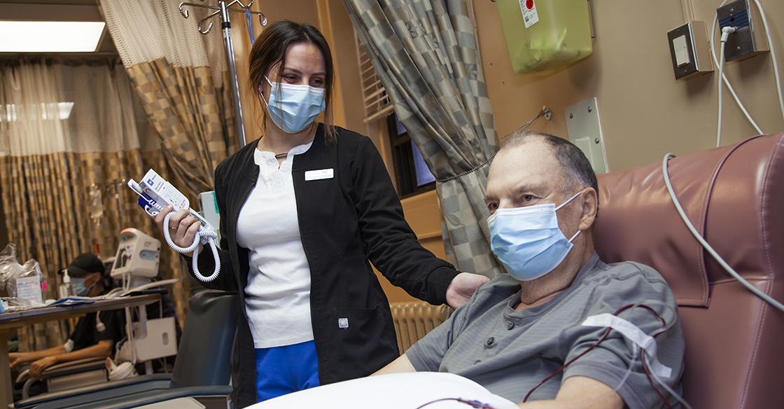 A QEII nurse stands next to an older male patient with her hand on his shoulder laying in bed receiving chemotherapy therapy treatment