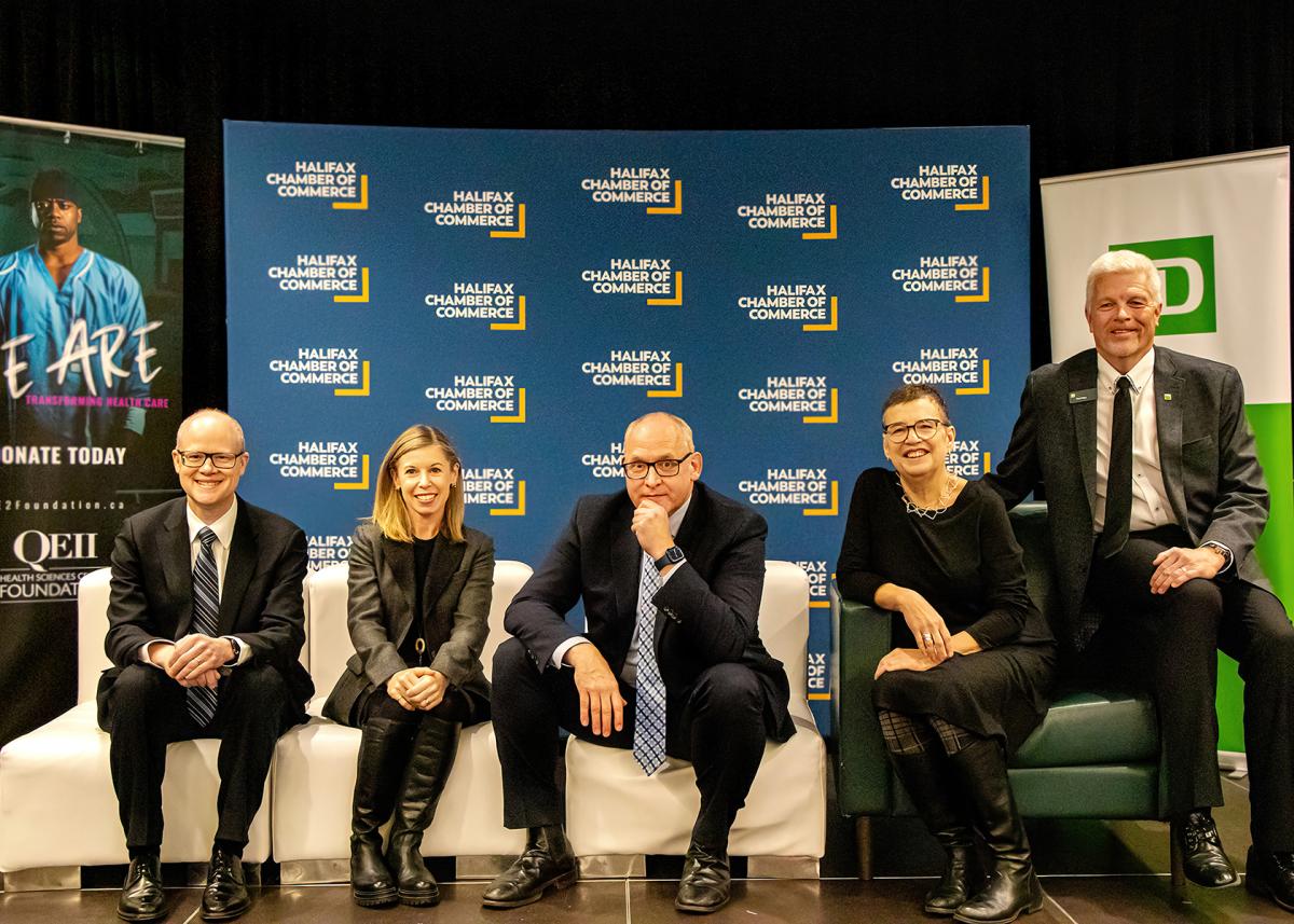 Five people wearing business attire sitting on a stage and smiling at the camera.