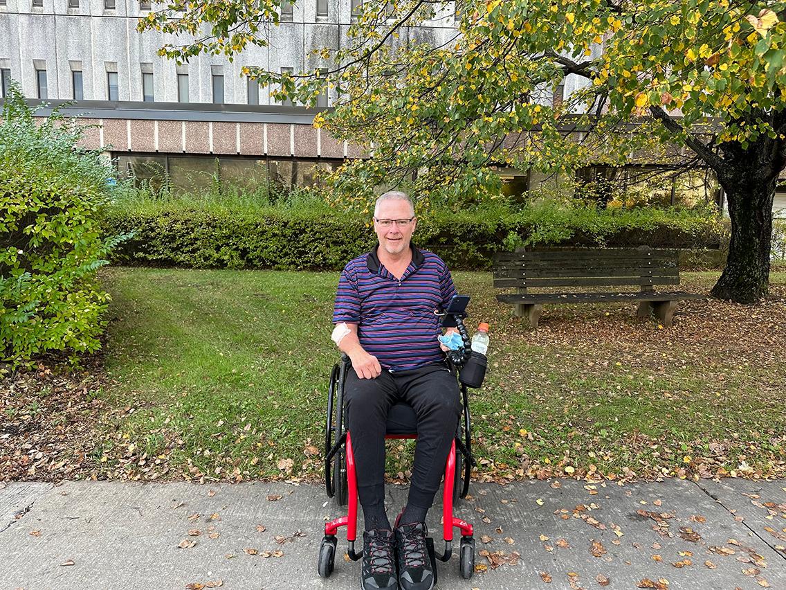 A man sits in a wheelchair in front of a brick building, surrounded by foliage.