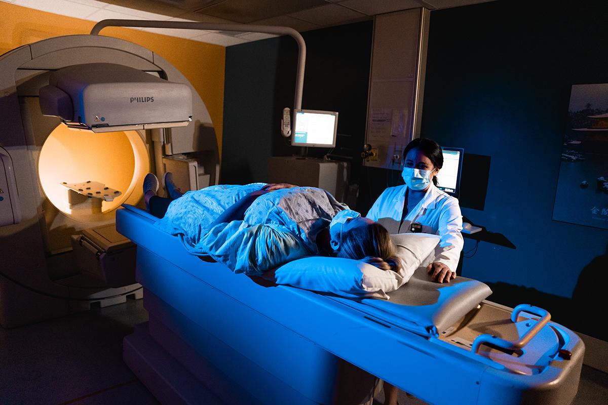 A female patient is laying down, preparing to undergo a medical scan. A female healthcare professional stands close to the patient. 