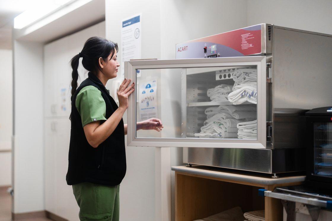 A nurse reaches into a machine that holds warm blankets.