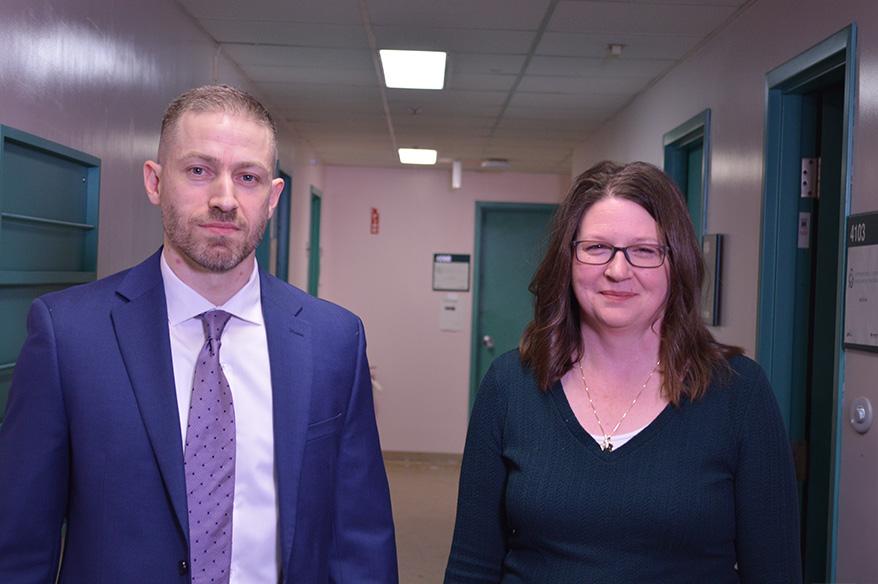 Two people, one wearing a suit, stand in a hallway, smiling.