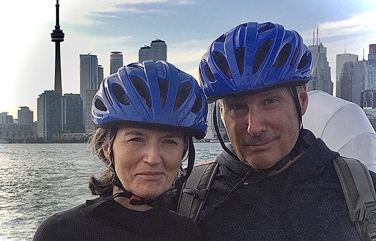 A couple wearing bicycle helmets pose in front of the Toronto skyline.