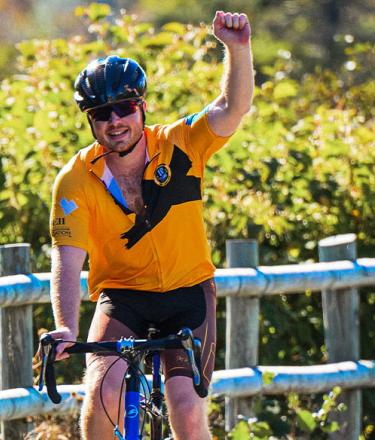 Rider in bright golden yellow jersey cycles along green trees with his fist raised in the air