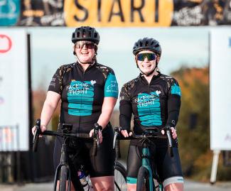 Two cyclists pose in helmets and cycling jerseys in front of a large start line banner
