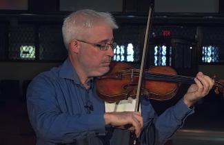 Renaud plays a violin with a church background. 
