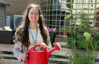A woman with long brown hair stands smiling outdoors in a garden. In the background is a building with lots of windows. She is surrounded by flowers and plants and holds a watering can.