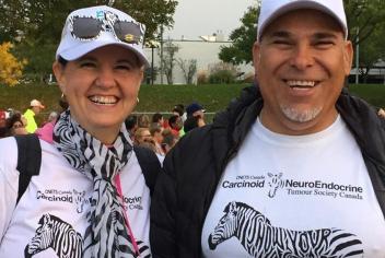 Jackie stands alongside her husband, Joe, at a busy, outdoor event. Both are smiling and wearing white t-shirts with zebra graphics