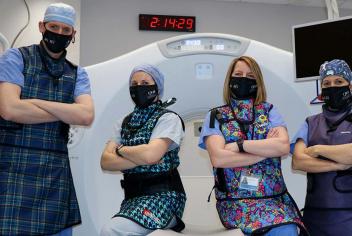 Group of four IR team members wear black We Are masks, while standing in front of large, white CT machine