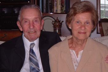 Elderly couple sit together on white sofa; husband's hand is on his wife's shoulder