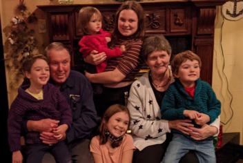 Elderly couple surrounded by five young grandkids, all smiling and hugging