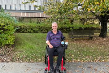 A man sits in a wheelchair in front of a brick building, surrounded by foliage. 