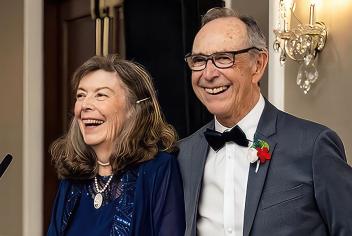 Couple speaking into microphone at son's wedding, wearing fancy clothes and beaming with pride