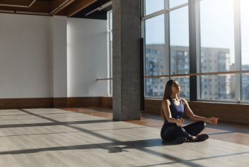 A woman sits in a yoga pose, facing large glass windows. 